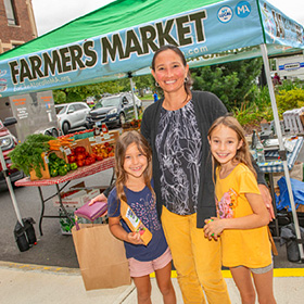 woman with two young girls standing in front of a farmers market booth