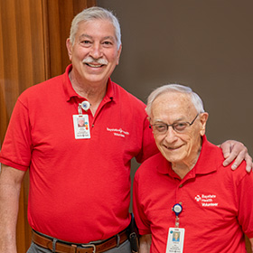 Two senior gentlemen volunteers, arm in arm, smiling.