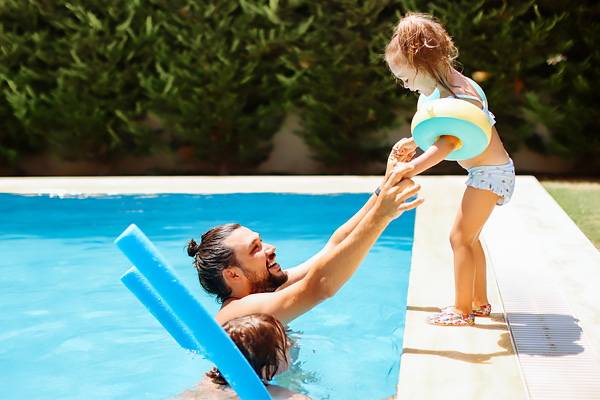 a dark-haired man waits in a pool to safely catch a little girl, standing on the side of the pool wearing personal floatation devices