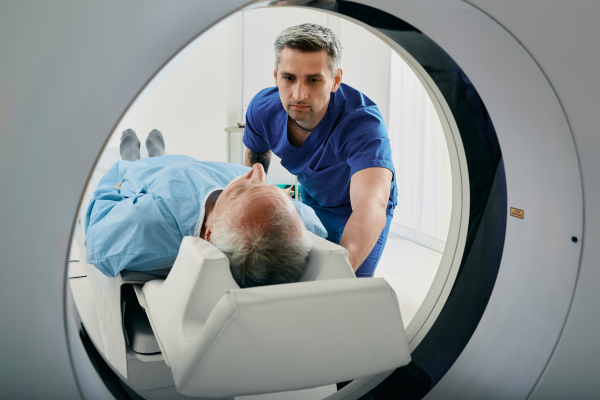 A man in blue scrubs guides a patient wearing a blue robe into a CT scanner.