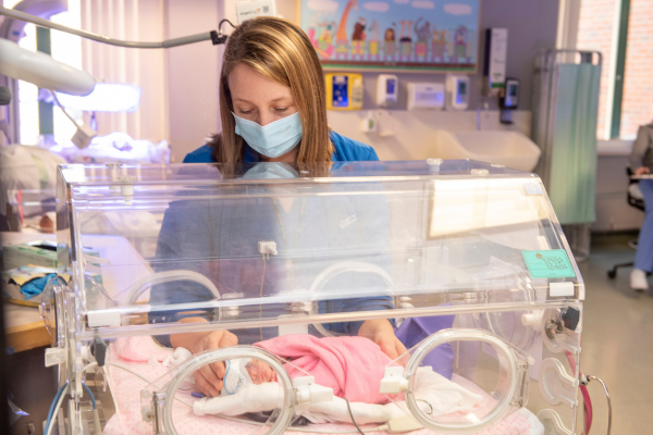 A nurse with a blue mask looks in on a baby in a Neonatal Intensive Care Unit bed at Baystate Medical Center.
