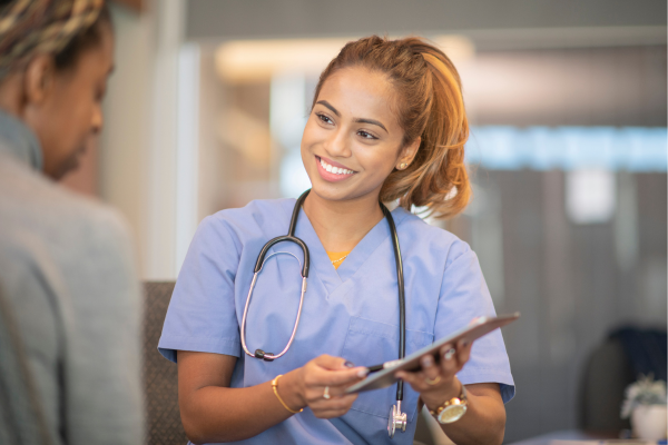 A woman in blue scrubs with a stethoscope shows another person something on a clipboard in a medical setting.