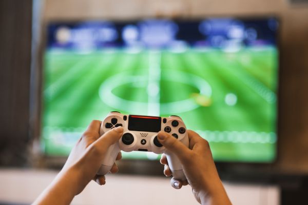 Hands hold up a video game remote control in front of a screen that has a video game on it.