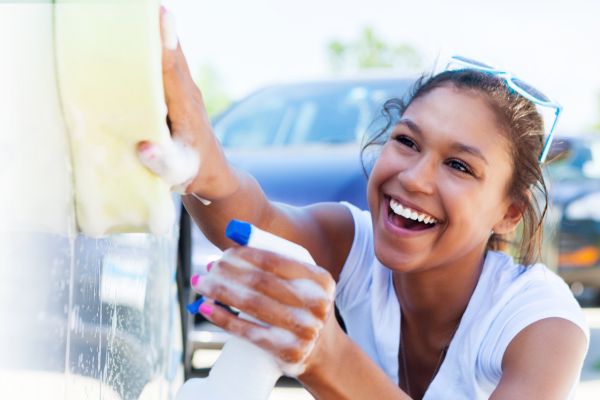 A smiling woman holds a sponge and spray bottle as she washes a car.
