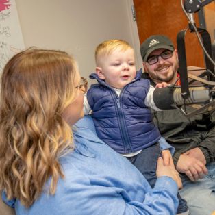 A woman in a blue shirt holds a young boy wearing a  blue vest with blond hair in front of a radio microphone. A man smiles behind the woman and boy.