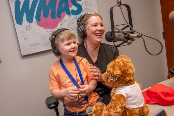 A young boy in an orange shirt with blonde hair wearing headphones sits with a woman with blond hair and a black shirt, also wearing headphones, in front of a radio station microphone.