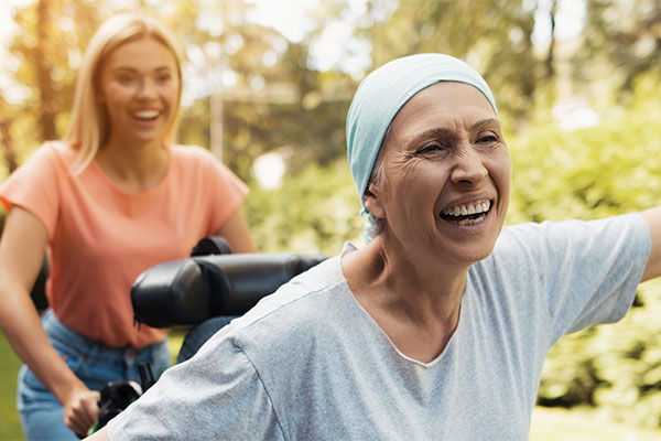 A patient smiles and outstretches her arms while a young person pushes her in a wheelchair.
