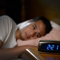 Person laying on bed, resting head on pillow while staring at alarm clock that reads 2:21 a.m.