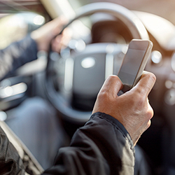 person sitting in vehicle with one hand placed on steering wheel and one hand holding cell phone with thumb on phone screen.