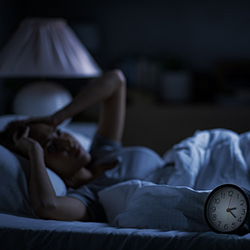 person laying next to alarm clock on a bed in dark room with open eyes and both hands on forehead.
