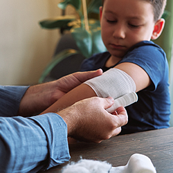 Young boy sitting down while watching someone his wrap arm in gauze bandage