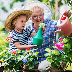 A toddler in a striped shirt with an older woman wearing glasses and a plaid shirt watering their garden together