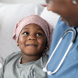 a child wearing a head covering and a hospital gown smiling up at a doctor wearing scrubs and smiling back