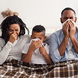 A family sitting on their couch, blowing their noses, covered in a blanket