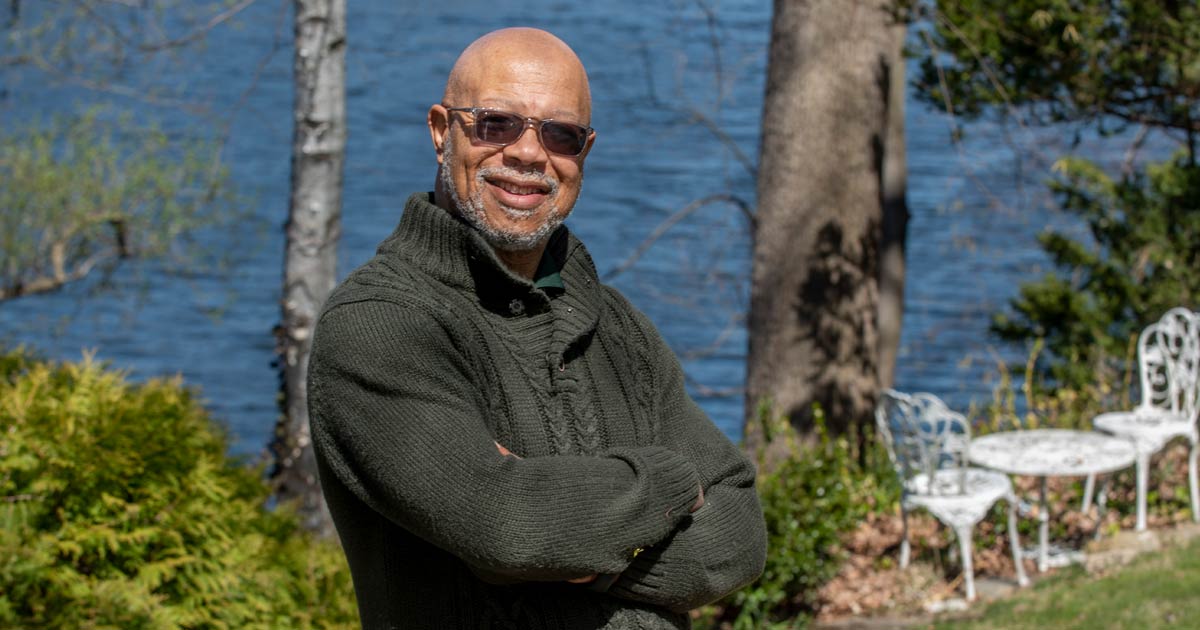 Dr. Frank Robinson wearing tinted sunglasses outside, smiling with arms crossed. Trees, white table and chair set, and body of water in background. 
