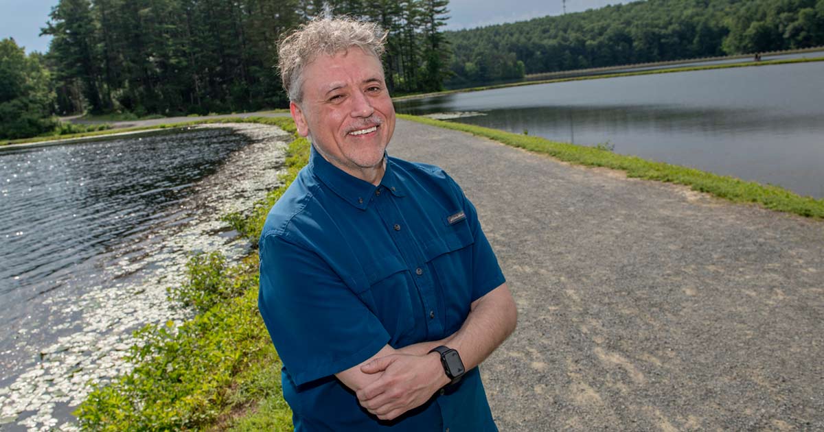 a man in a blue shirt smiling in front of a path and a body of water