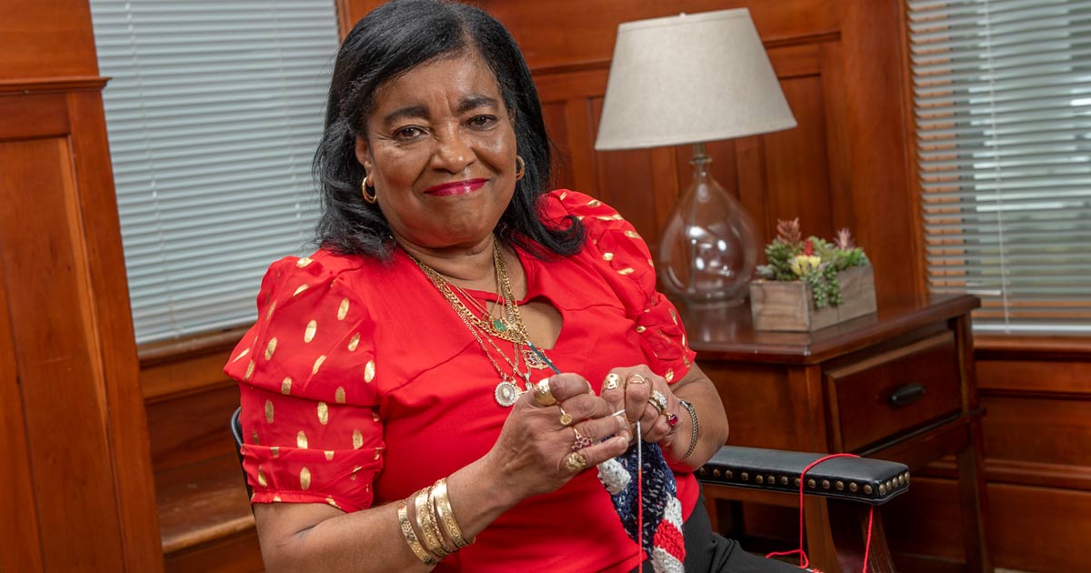 A Black woman in a red dress, crocheting and smiling