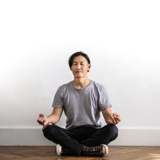 A man in a grey T-shirt and black pants sits with his eyes closed with a white wall behind him.