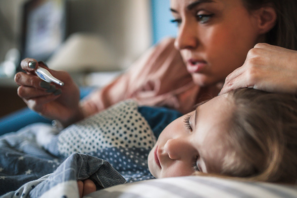 Mother holds thermometer and lays next to their child, who is resting their head on a pillow.