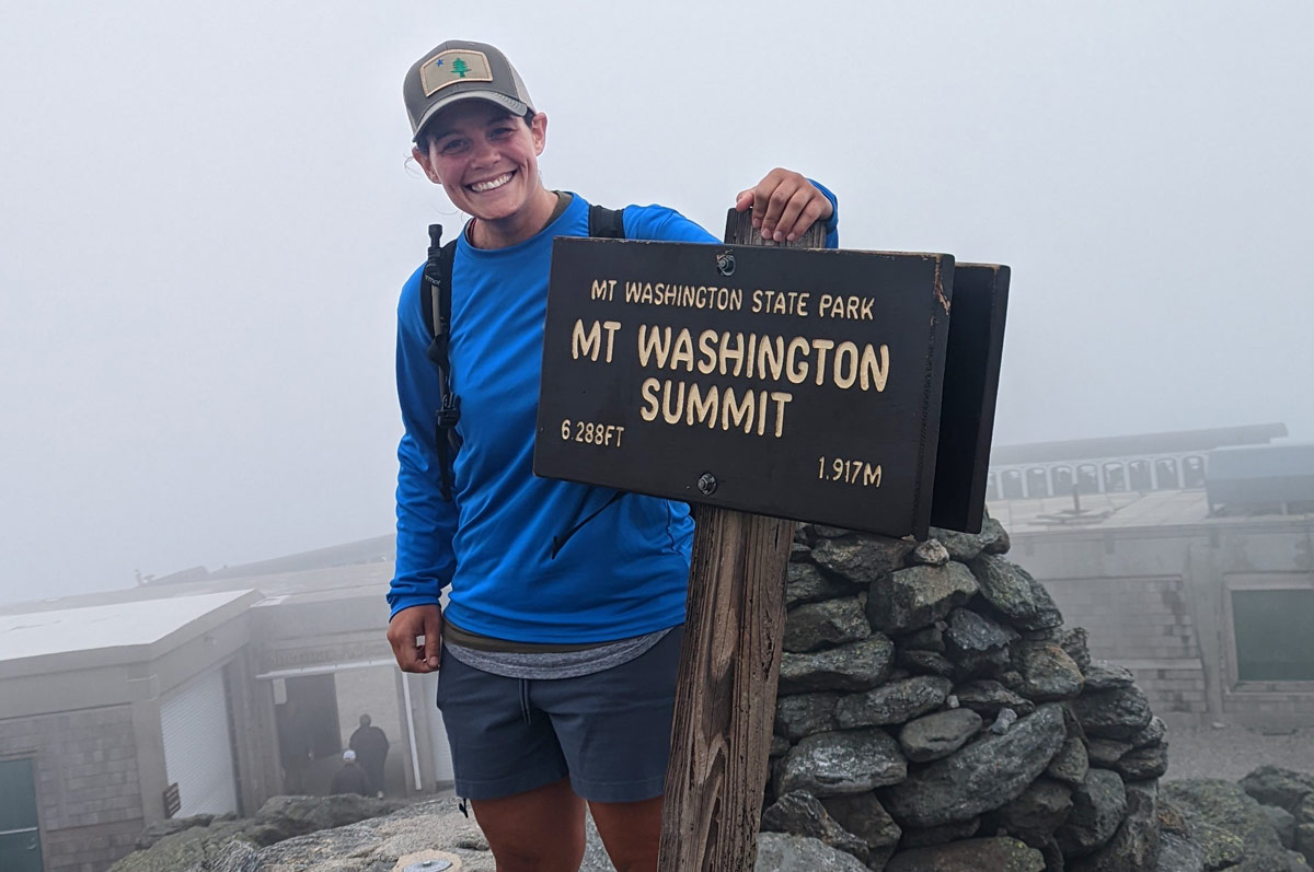 A smiling woman wearing bright blue standing with the Mt. Washington Summit sign on a foggy day