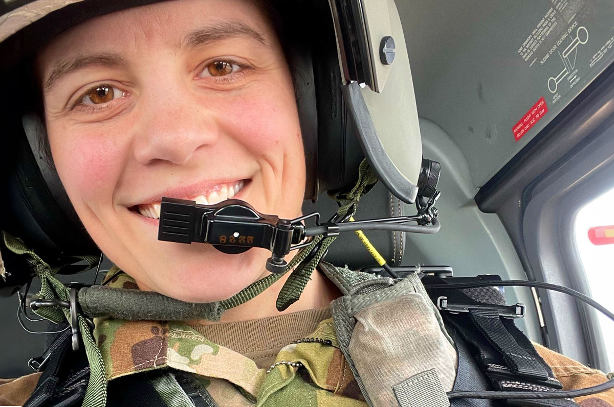 An army woman in a helicopter wearing her helmet and microphone, smiling at the camera