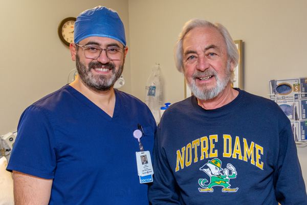 a male doctor with a beard and glasses in blue scrubs and a male patient with white hair and a beard smiling after a procedure