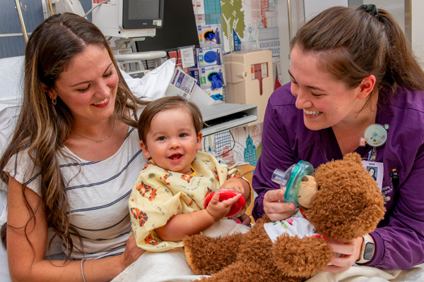 a pediatric hospital room scene with a smiling toddler, mom, and medical professional