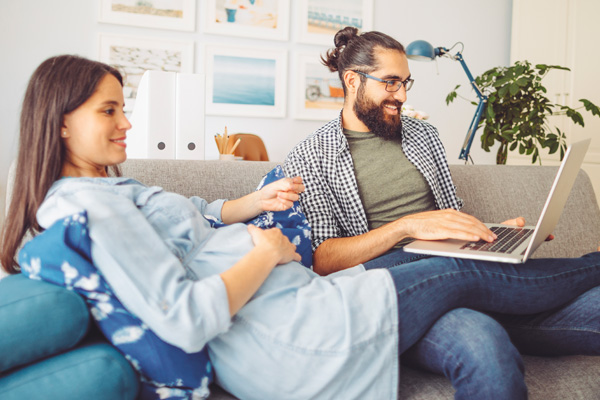 young pregnant couple together on the couch using a laptop computer