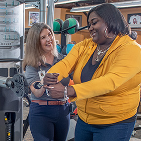 a rehabilitation staff member assists in a young woman’s therapy as she uses an upper-body weight-strengthening machine